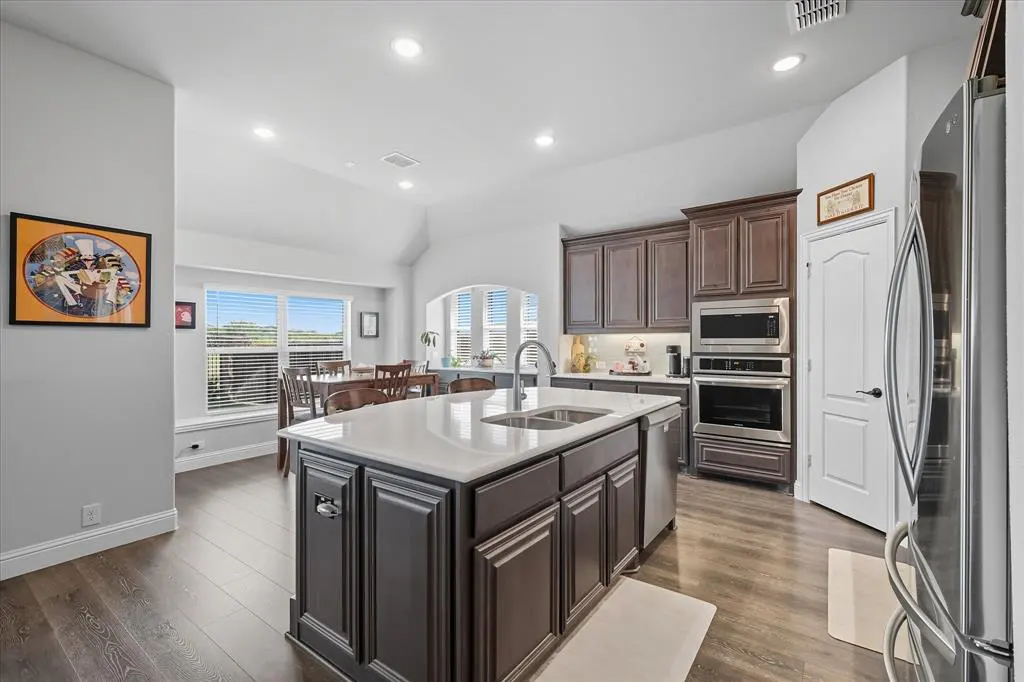 A bright kitchen with dark wood cabinets, stainless steel appliances, and a white countertop island. A dining table is visible in the background.