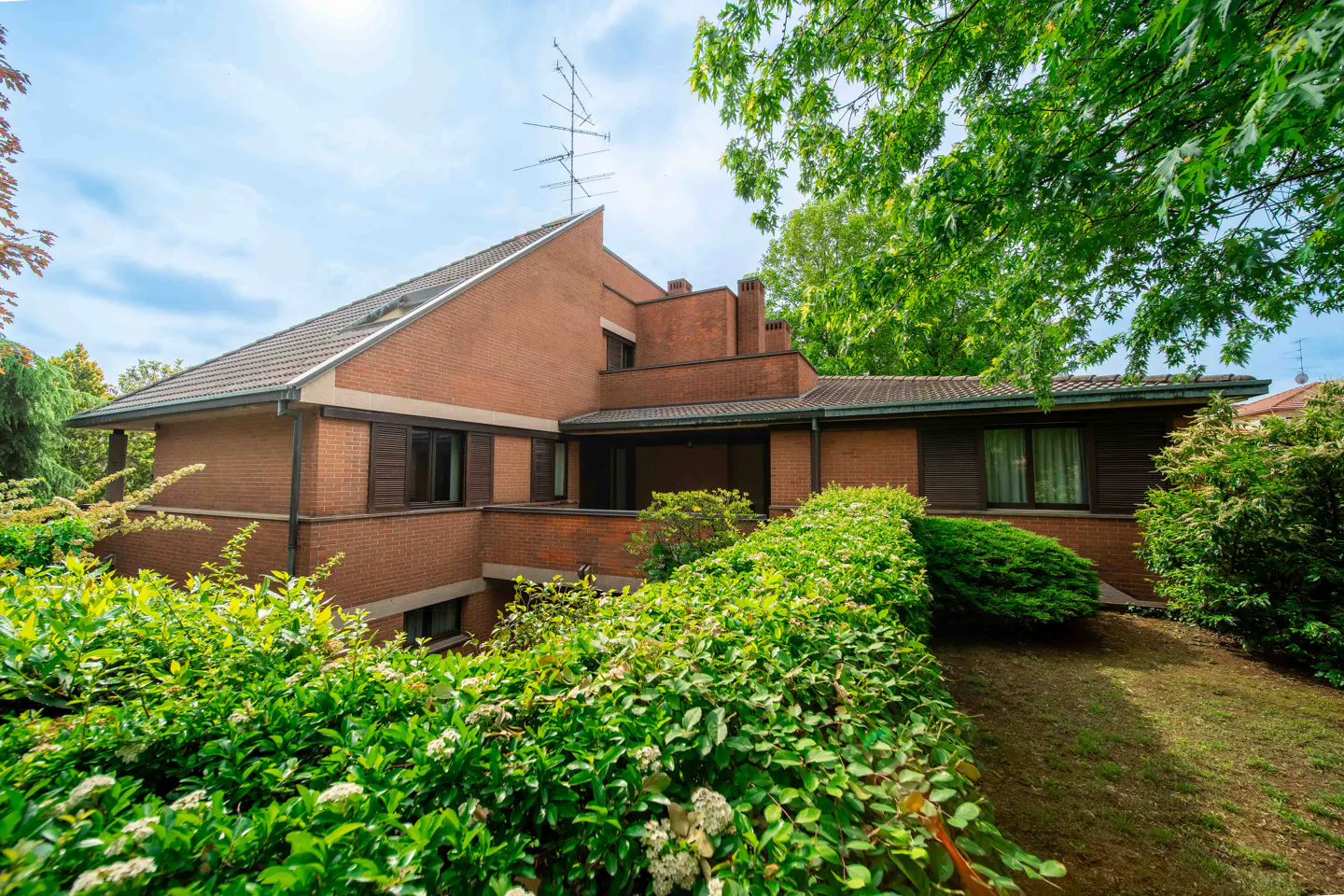 Exterior view of a brick house with brown trim, surrounded by green bushes and trees under a blue sky.