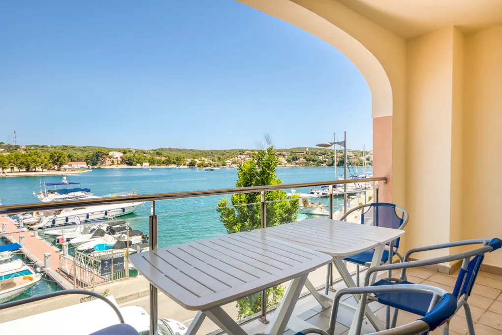 Balcony view of a marina with boats. White table and blue chairs on the balcony. Clear blue sky.