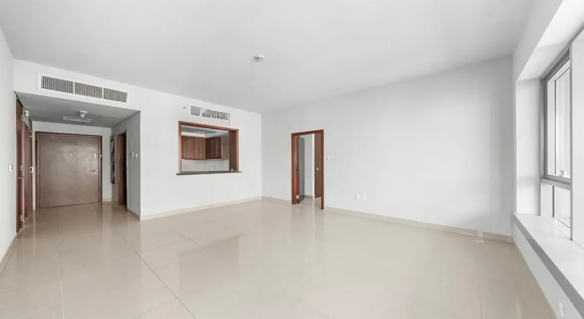 Empty apartment with white walls, beige tile floor, and a pass-through to a kitchen with wood cabinets. A window is on the right.