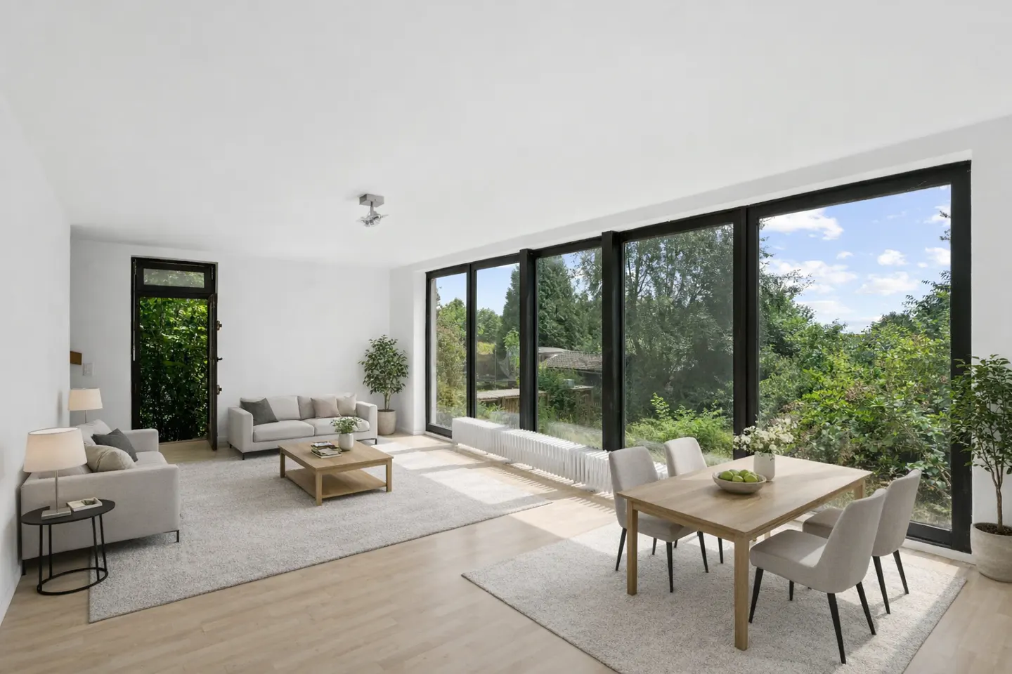 Bright, modern living and dining area with large windows overlooking lush greenery. Light wood floors, neutral furniture, and a cozy rug create an inviting space.