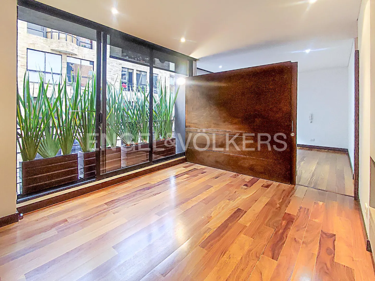 A bright room with wood floors, a large window with plants, and a dark wood door.