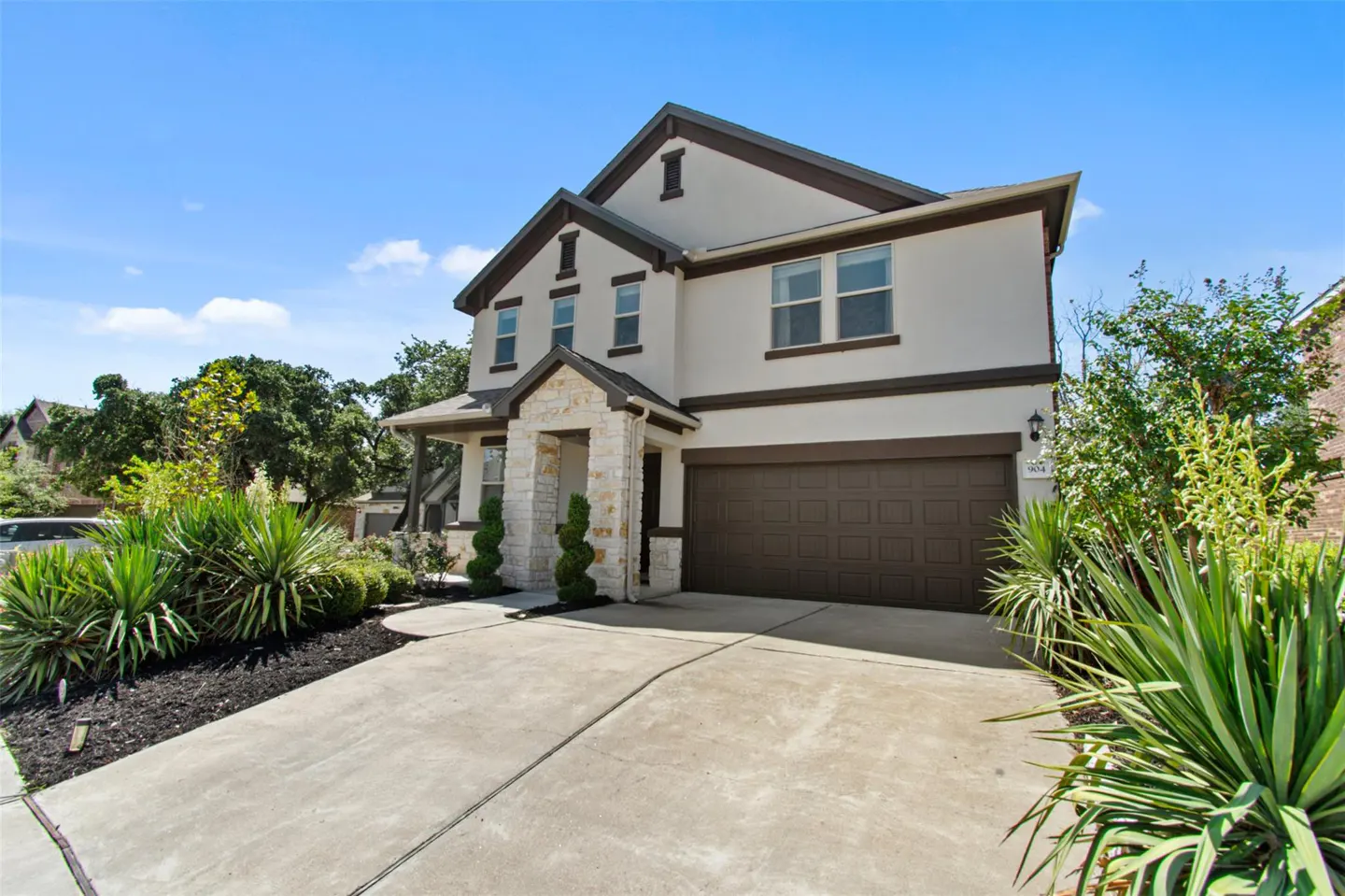 Two-story house with a brown garage door, stone accents, and green landscaping under a blue sky.