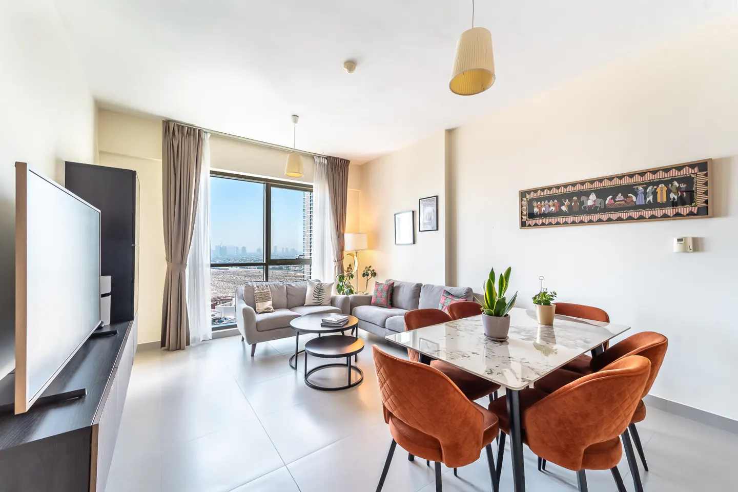 A bright living room with gray sofas, a marble table with orange chairs, and a large window with city views.