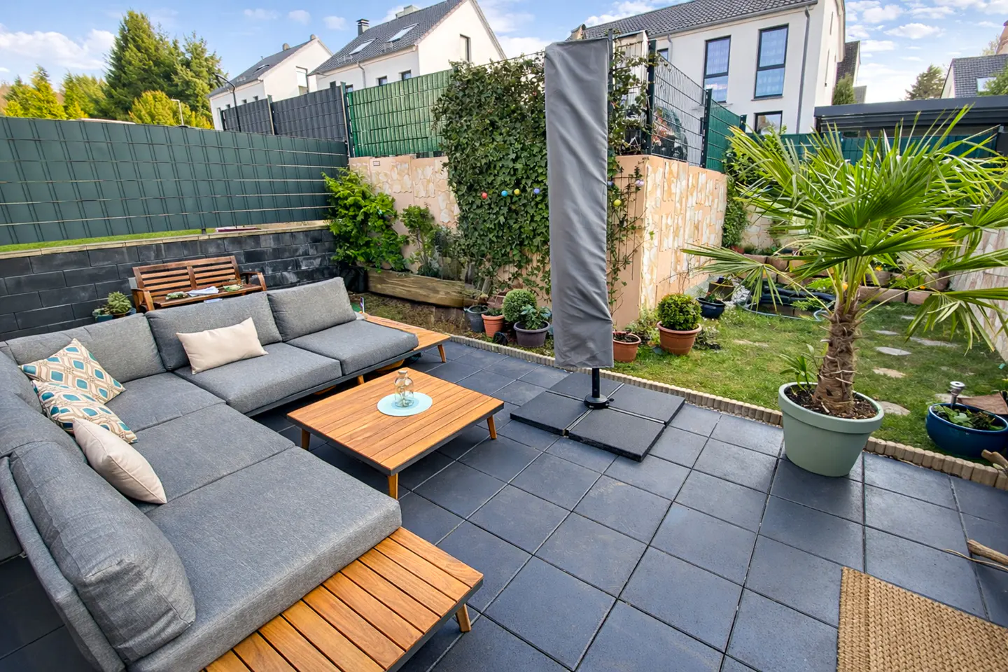 A backyard patio with a gray sectional sofa, wooden coffee table, and a covered umbrella. A palm tree sits in a pot on the lawn.