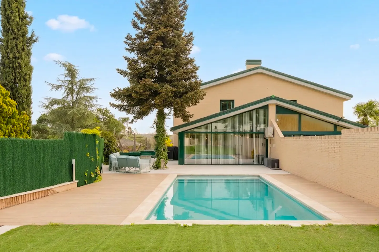 Backyard view of a beige house with a pool, wooden deck, green trim, and a tall tree. Blue sky above.
