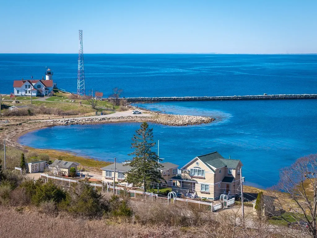 Coastal view of a lighthouse, pier, and houses against a blue sky and ocean backdrop.