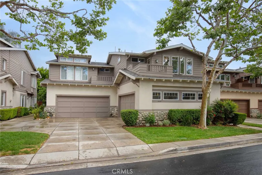 Two-story tan house with a two-car garage, stone accents, and green bushes. Trees frame the house against a blue sky.