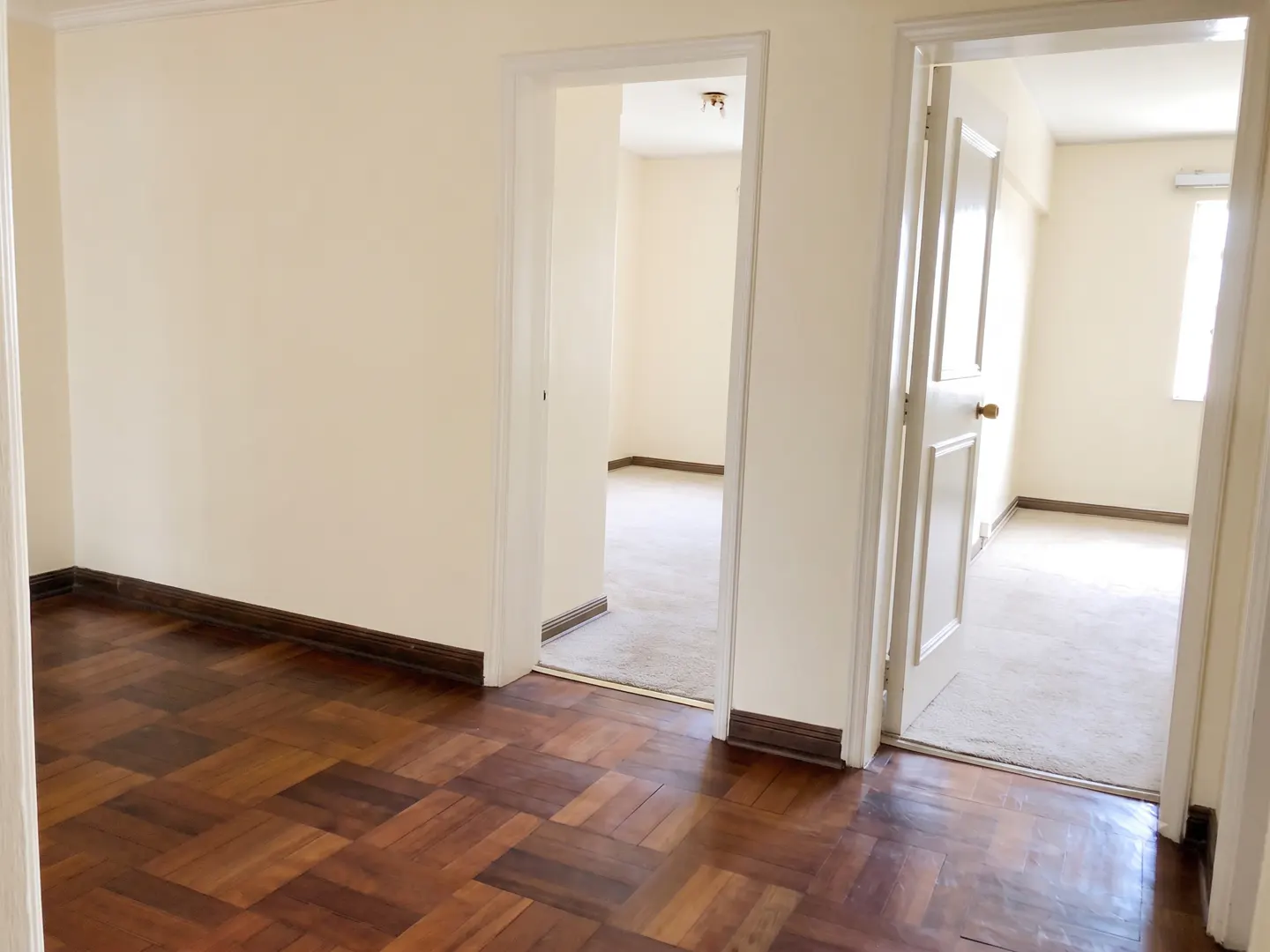 A room with dark parquet wood floors and cream walls, featuring two doorways leading to carpeted rooms with natural light.