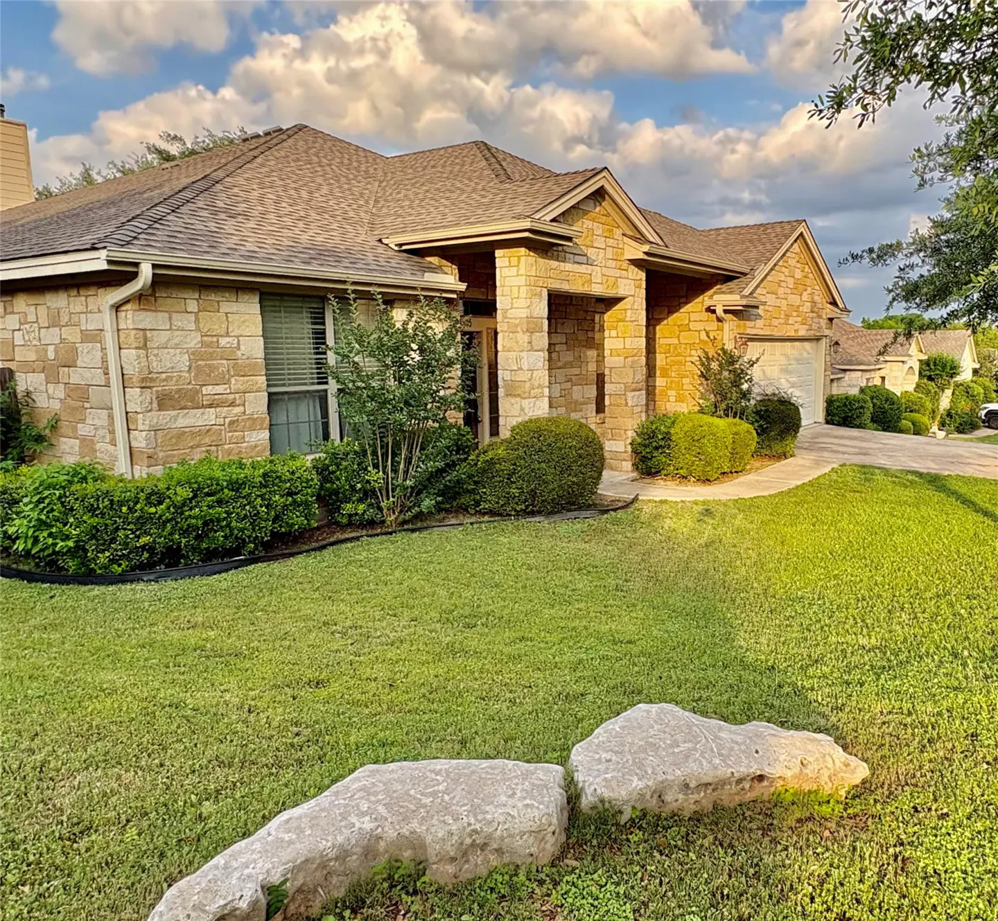 A tan stone house with a brown roof sits on a green lawn under a cloudy sky. Two large rocks are in the foreground.