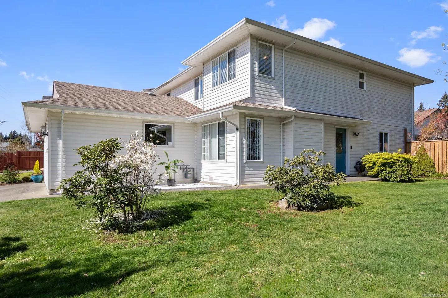 Two-story house with white siding, brown roof, and a blue front door, surrounded by green lawn and bushes.