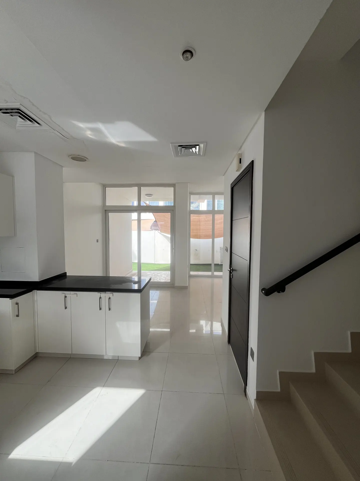 Bright, modern interior with white cabinets, black countertops, and tile floors. Sliding glass doors lead to a green lawn. Stairs with a black railing are visible.