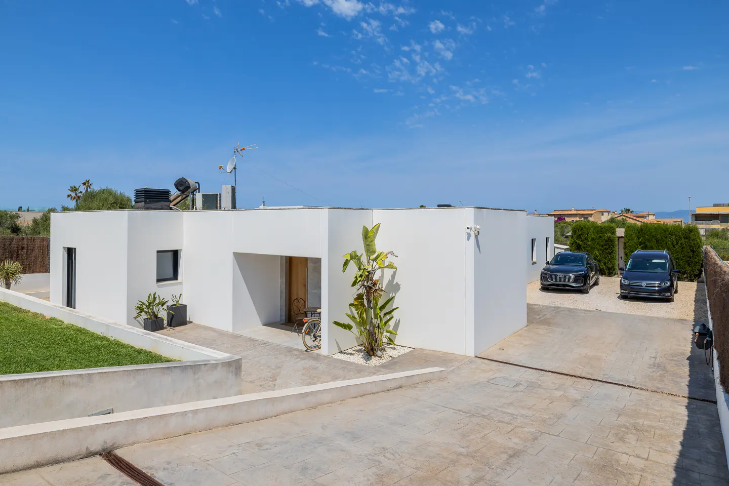 Modern white flat-roofed house with a paved driveway, two parked cars, a small garden, and a bright blue sky.