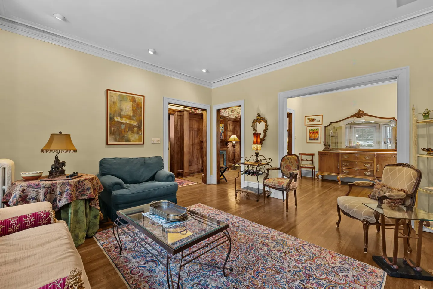 A living room with hardwood floors, a blue couch, a glass coffee table, and a colorful rug. There are doorways leading to other rooms.
