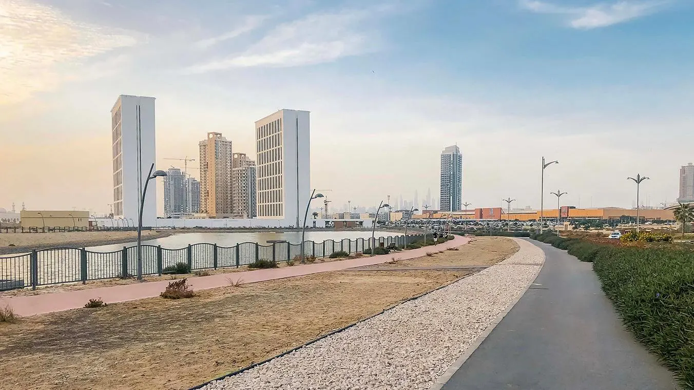 Cityscape view with walking path, lake, and modern buildings. Tall white buildings and other skyscrapers are visible against a blue sky.