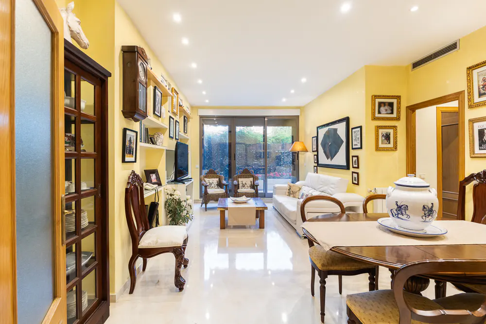 Bright, yellow living room with a dining table, white sofa, and glass doors to the outside. Walls are decorated with pictures and shelves.