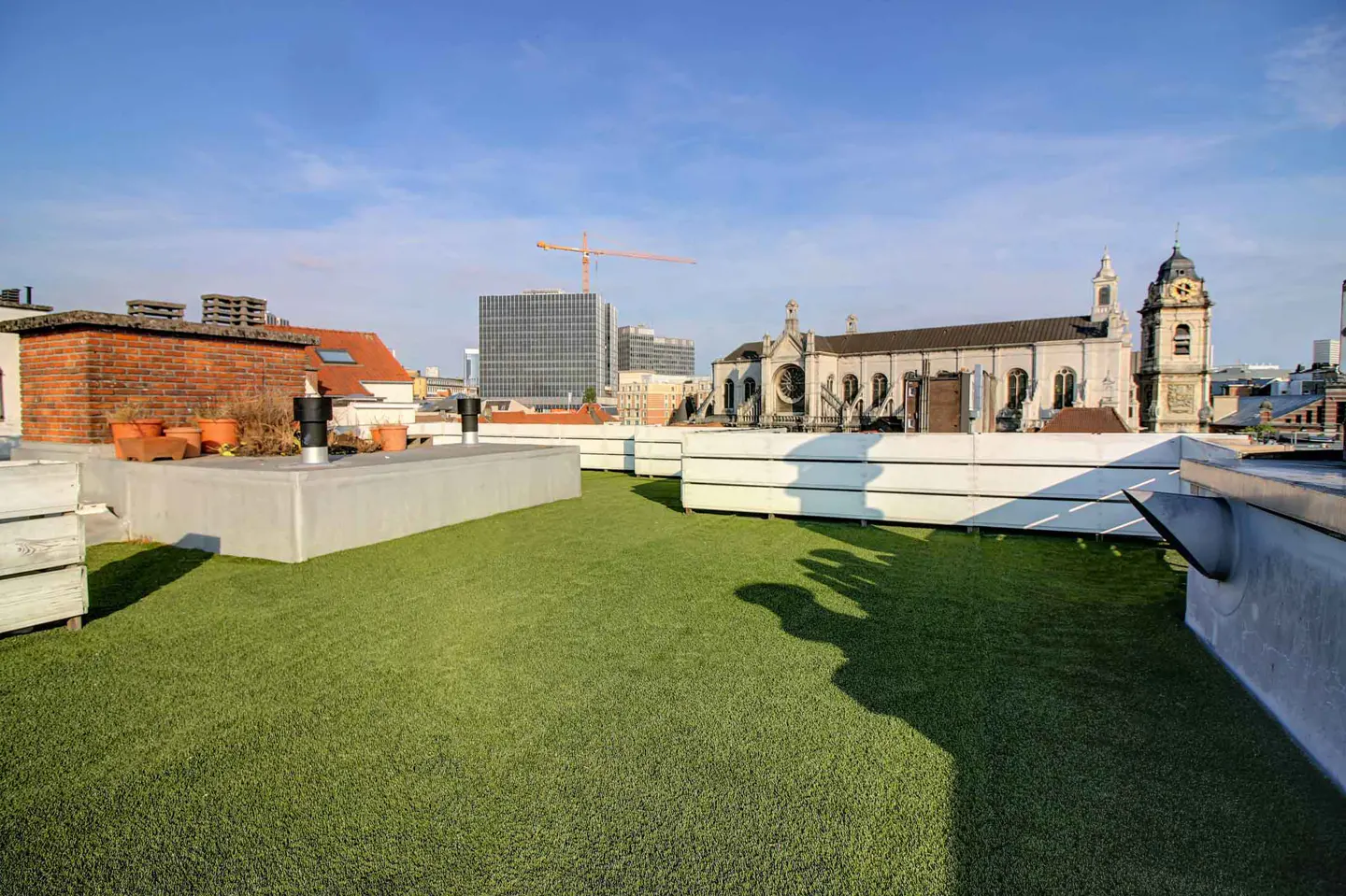 Rooftop view of Brussels, Belgium, with green turf, concrete planters, and a church and modern buildings in the background.