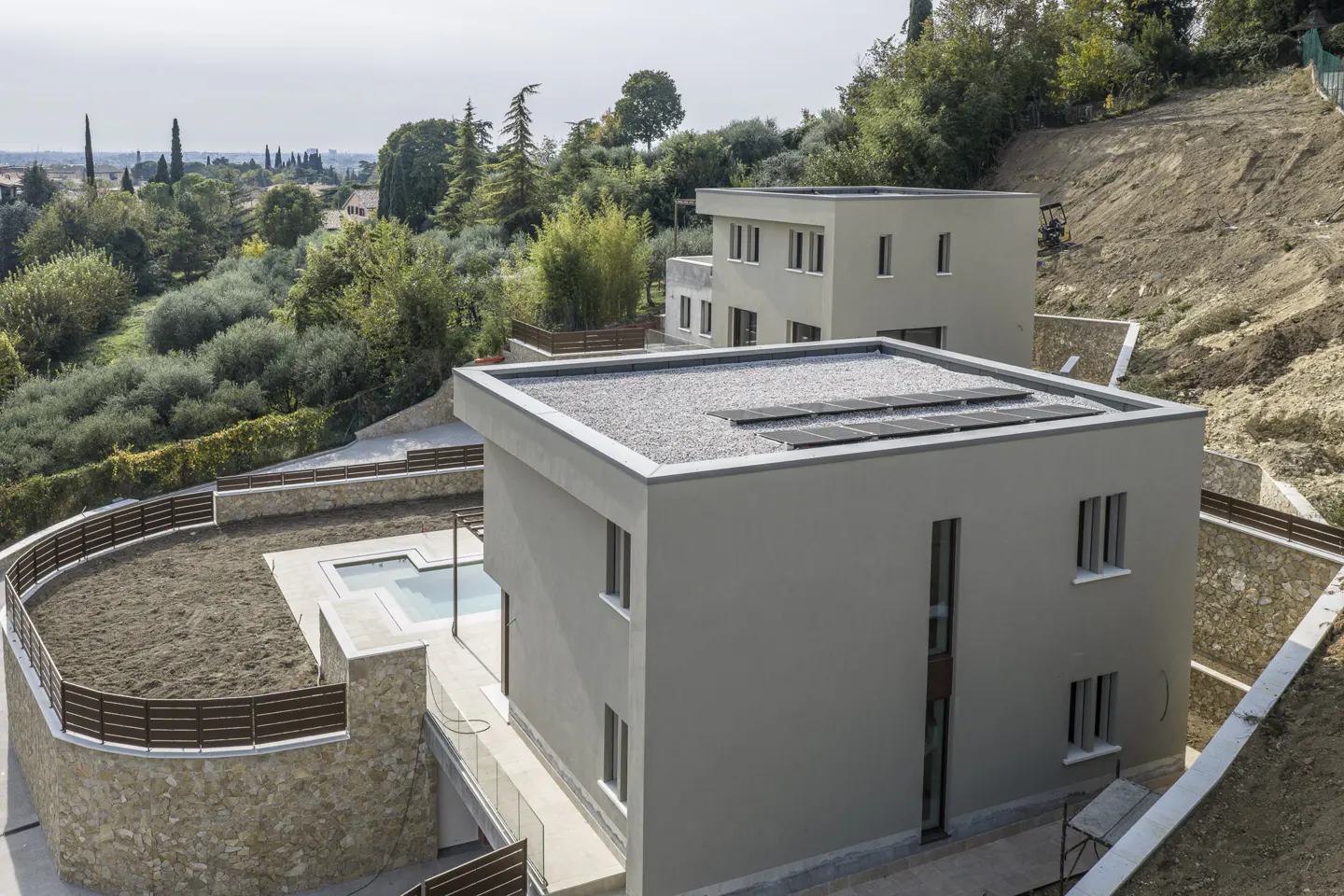 Exterior view of two modern, gray, flat-roofed homes with solar panels, a pool, and a stone retaining wall on a hillside.