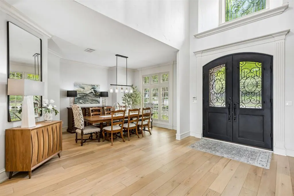 Bright foyer with wood floors, black iron double doors, and a dining area with a long wooden table and chairs. A wooden cabinet with a mirror and lamps is on the left.