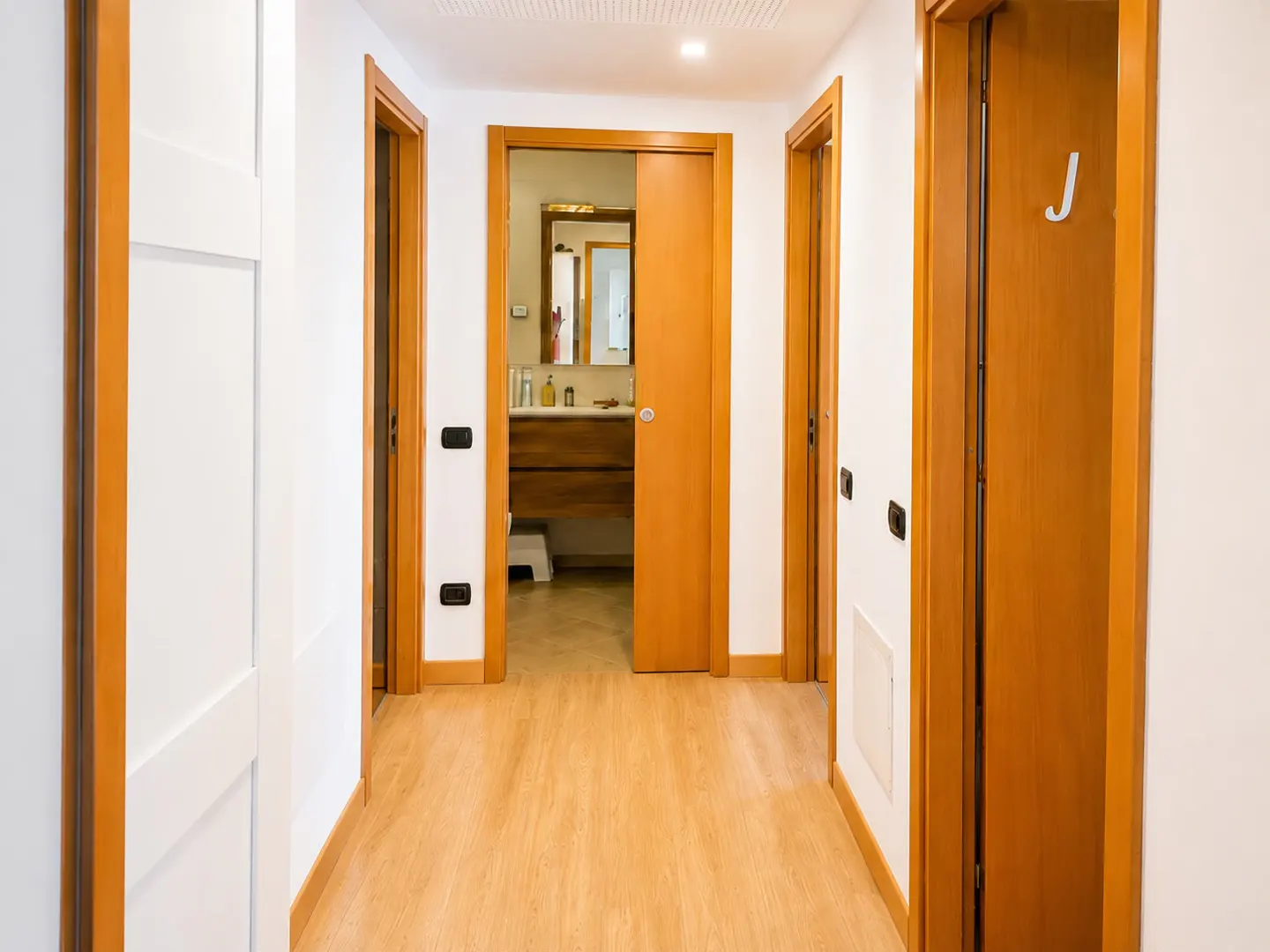 Bright hallway with light wood floors, white walls, and multiple wooden door frames leading to a bathroom.