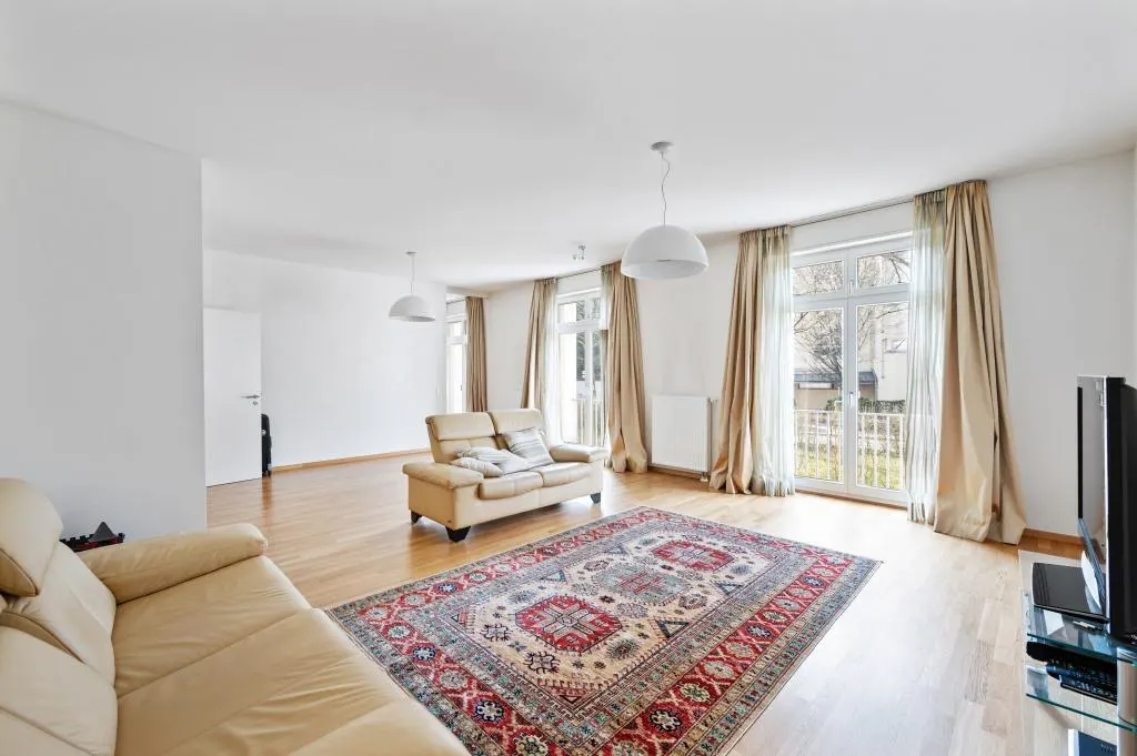 Bright living room with hardwood floors, beige sofas, and a red patterned rug. Large windows with beige curtains let in natural light.