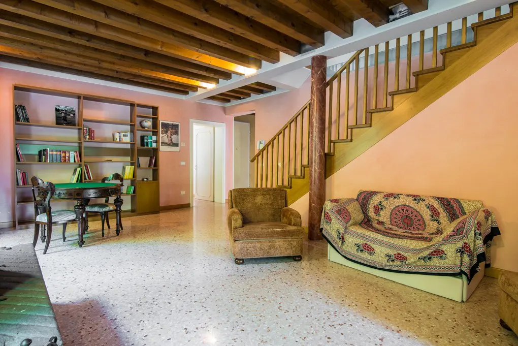 Living room with pink walls, wooden beamed ceiling, and terrazzo floor. A bookshelf, table, chairs, armchair, and sofa are visible.