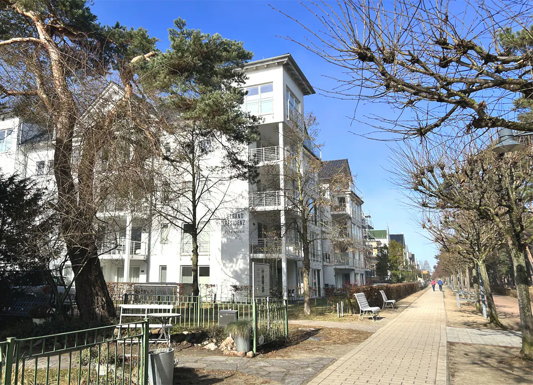 Exterior view of a white apartment building with balconies, trees, and a brick walkway on a sunny day.