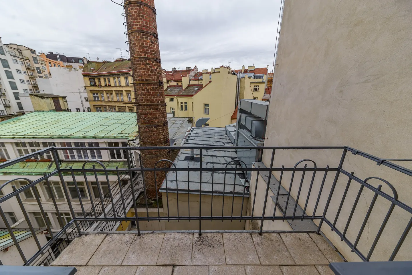 View from a balcony with a metal railing overlooking rooftops and buildings in Prague on a cloudy day.