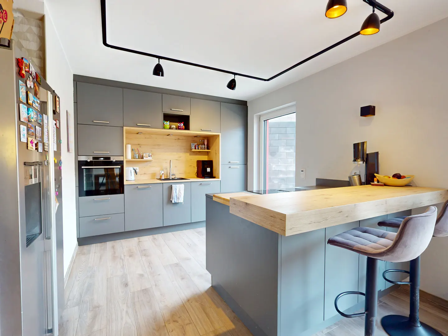 A modern kitchen with gray cabinets, a light wood countertop island with two gray stools, and black track lighting.