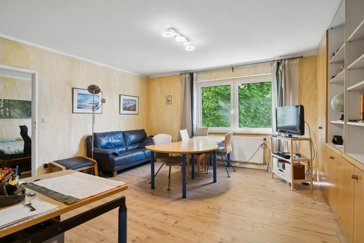 Living room with wood floors, yellow walls, blue leather sofa, round table, and a TV near a window.