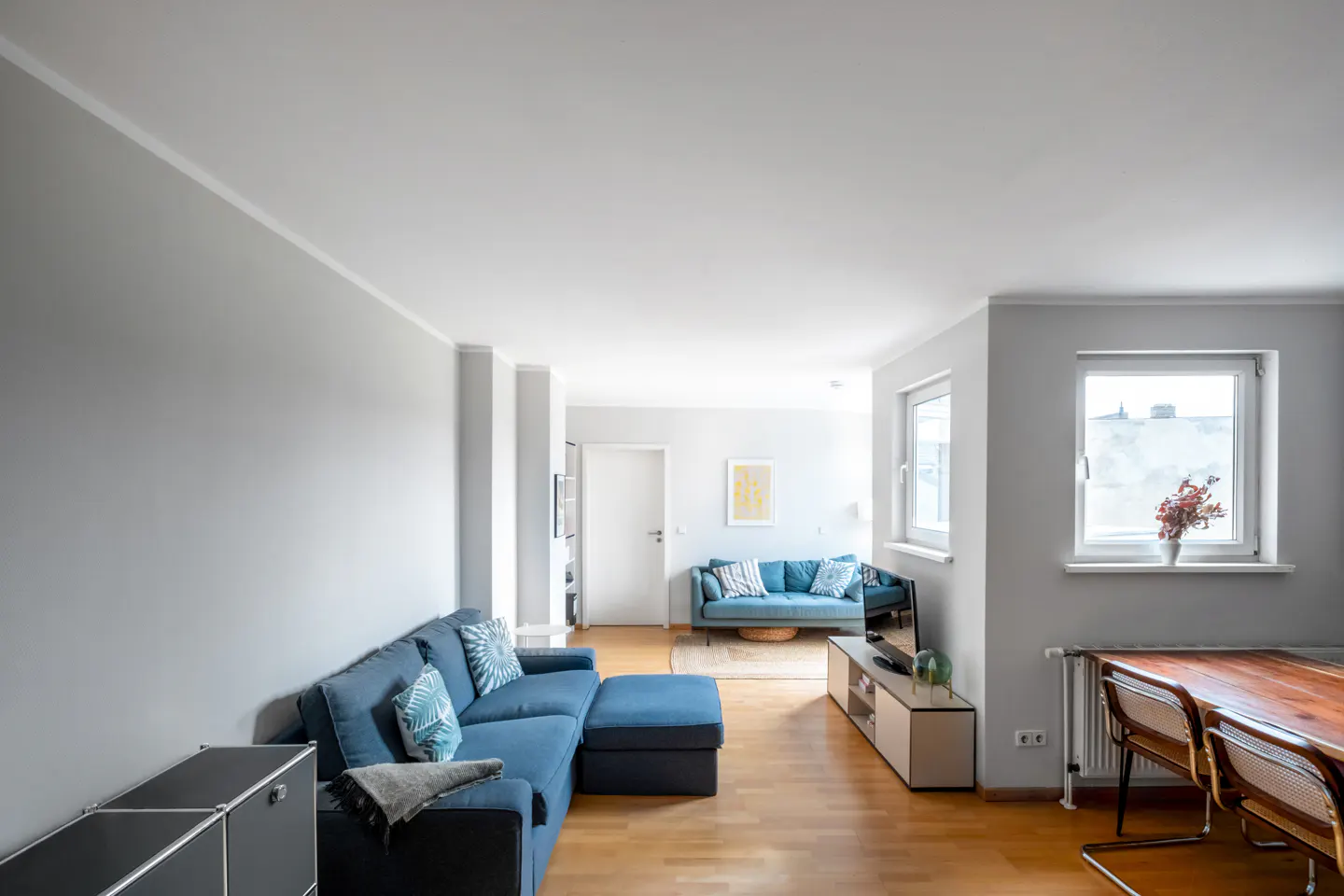 Bright living room with light gray walls, hardwood floors, two blue sofas, a wooden dining table, and large windows.