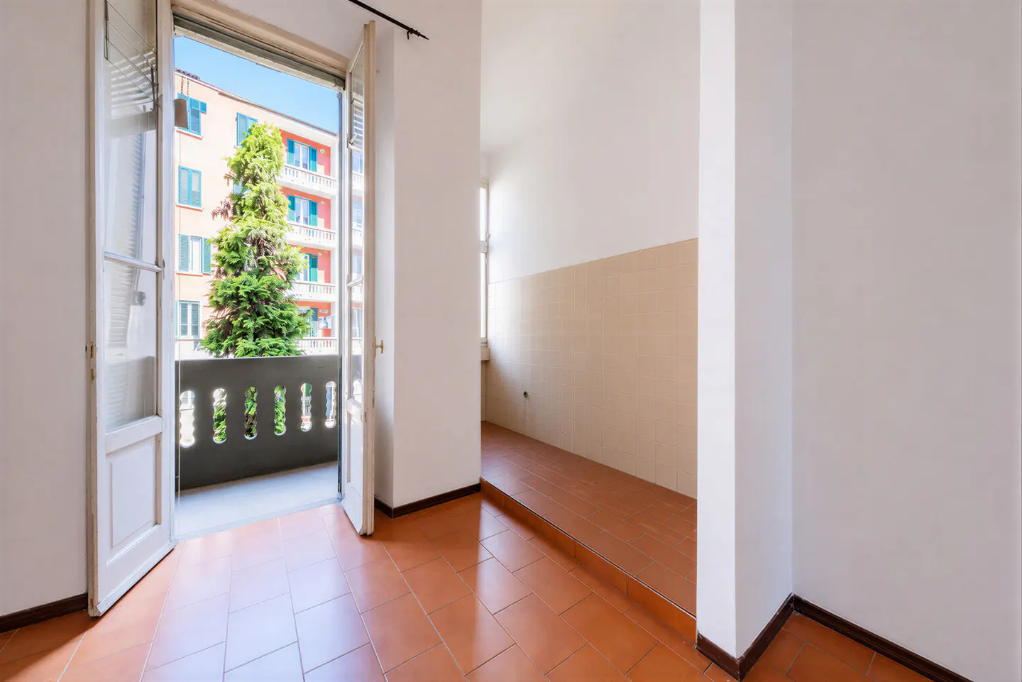 A bright room with terracotta tile floors and white walls, leading to an open balcony with a view of a tree and building.