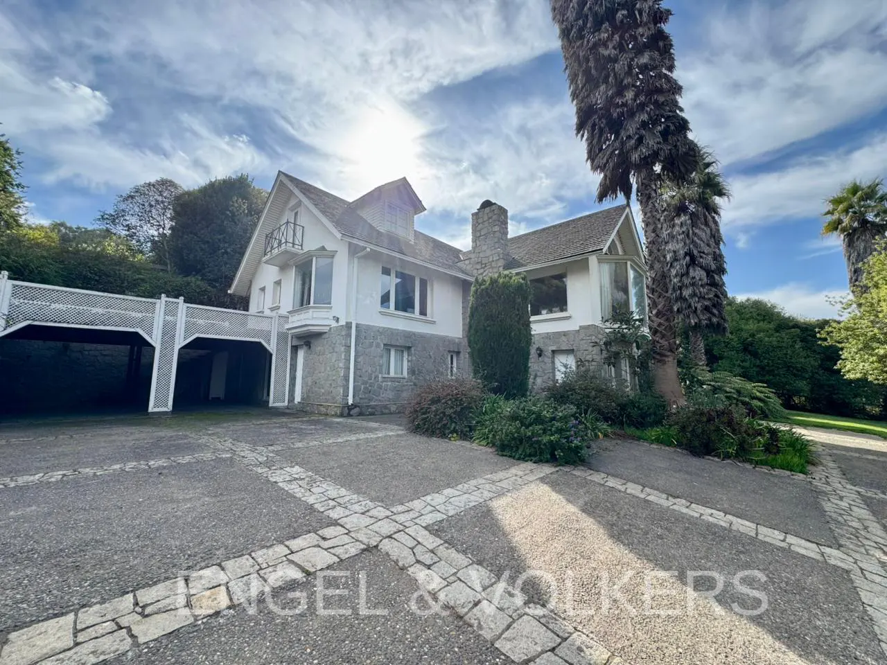 Exterior view of a two-story white house with stone accents, a covered carport, and a paved driveway. Palm trees and green foliage surround the property.