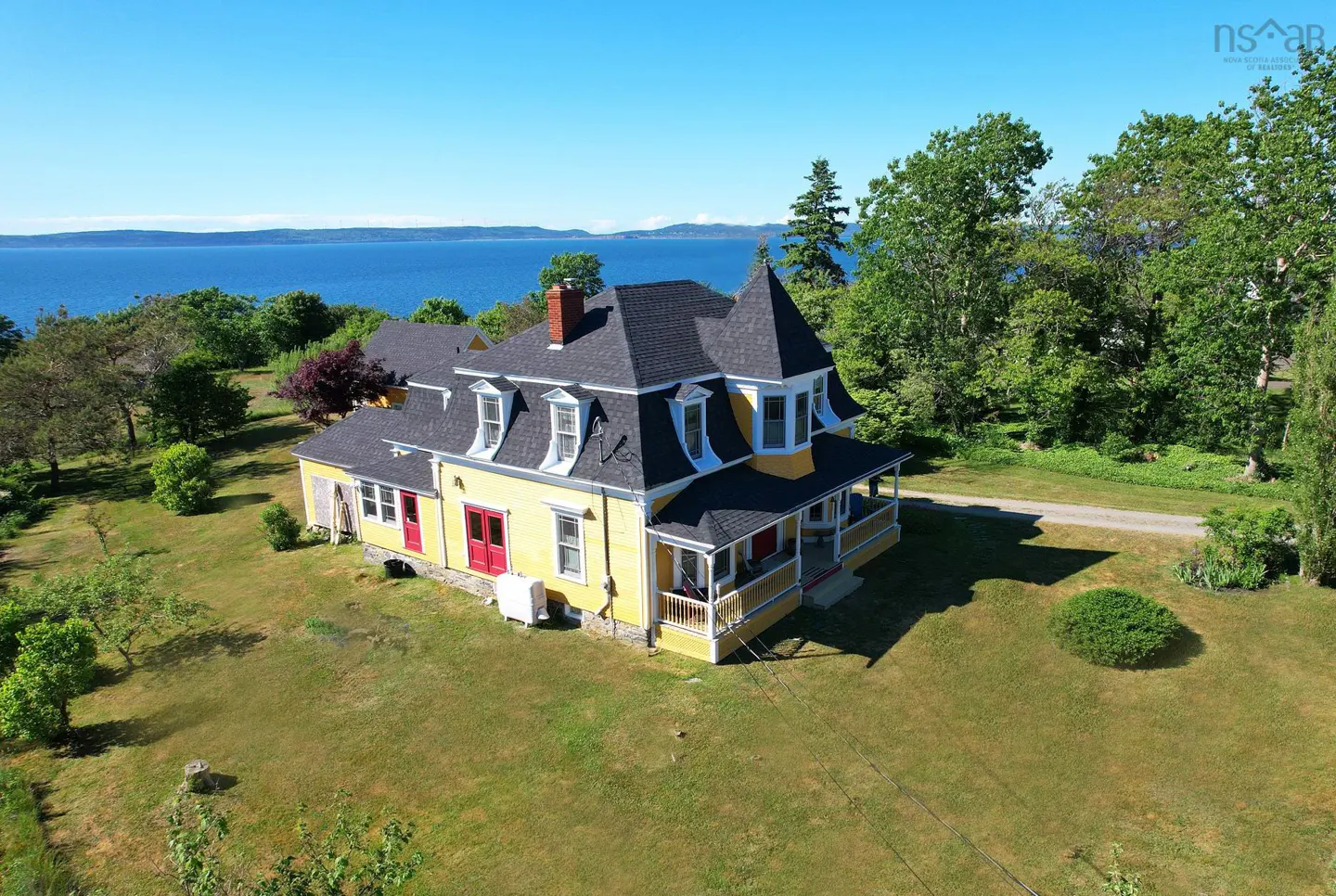 Aerial view of a yellow Victorian-style house with a dark roof and red doors, set on a grassy lot overlooking the ocean.