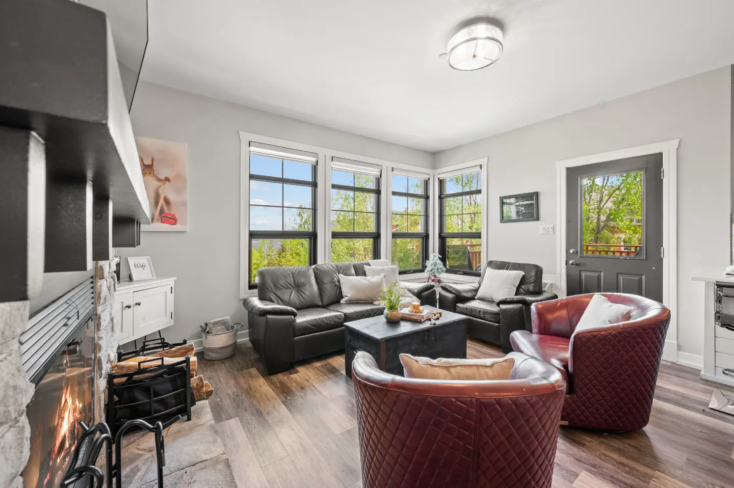 Living room with a stone fireplace, black leather sofa, two quilted maroon armchairs, and large windows with garden views.