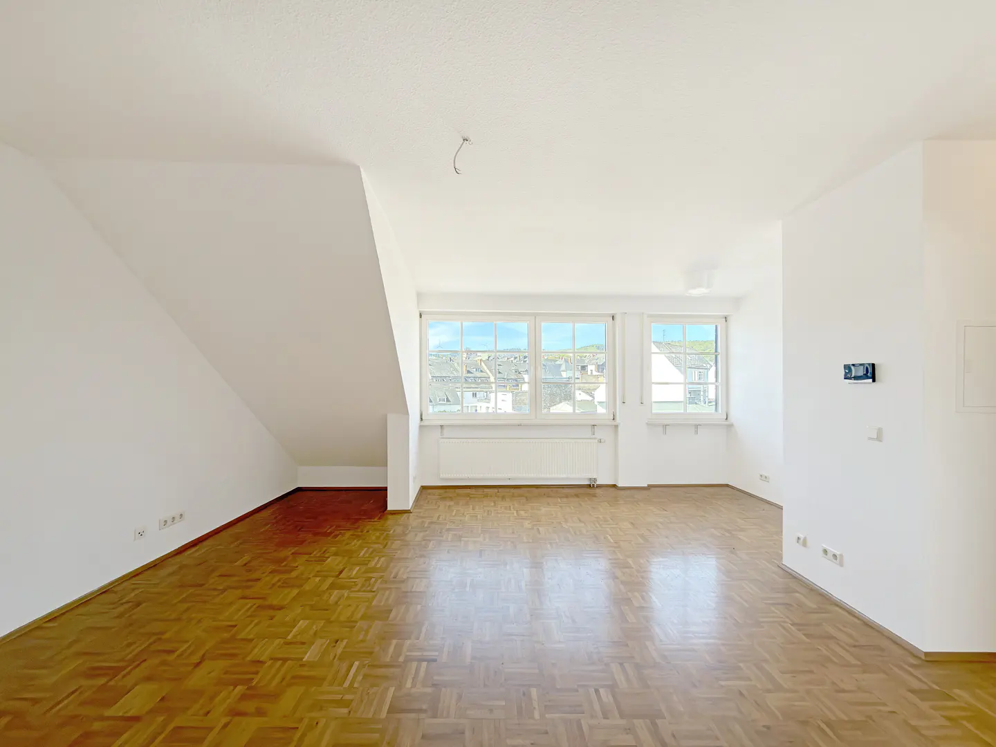 Bright, empty room with parquet floors, white walls, and two windows overlooking a town.