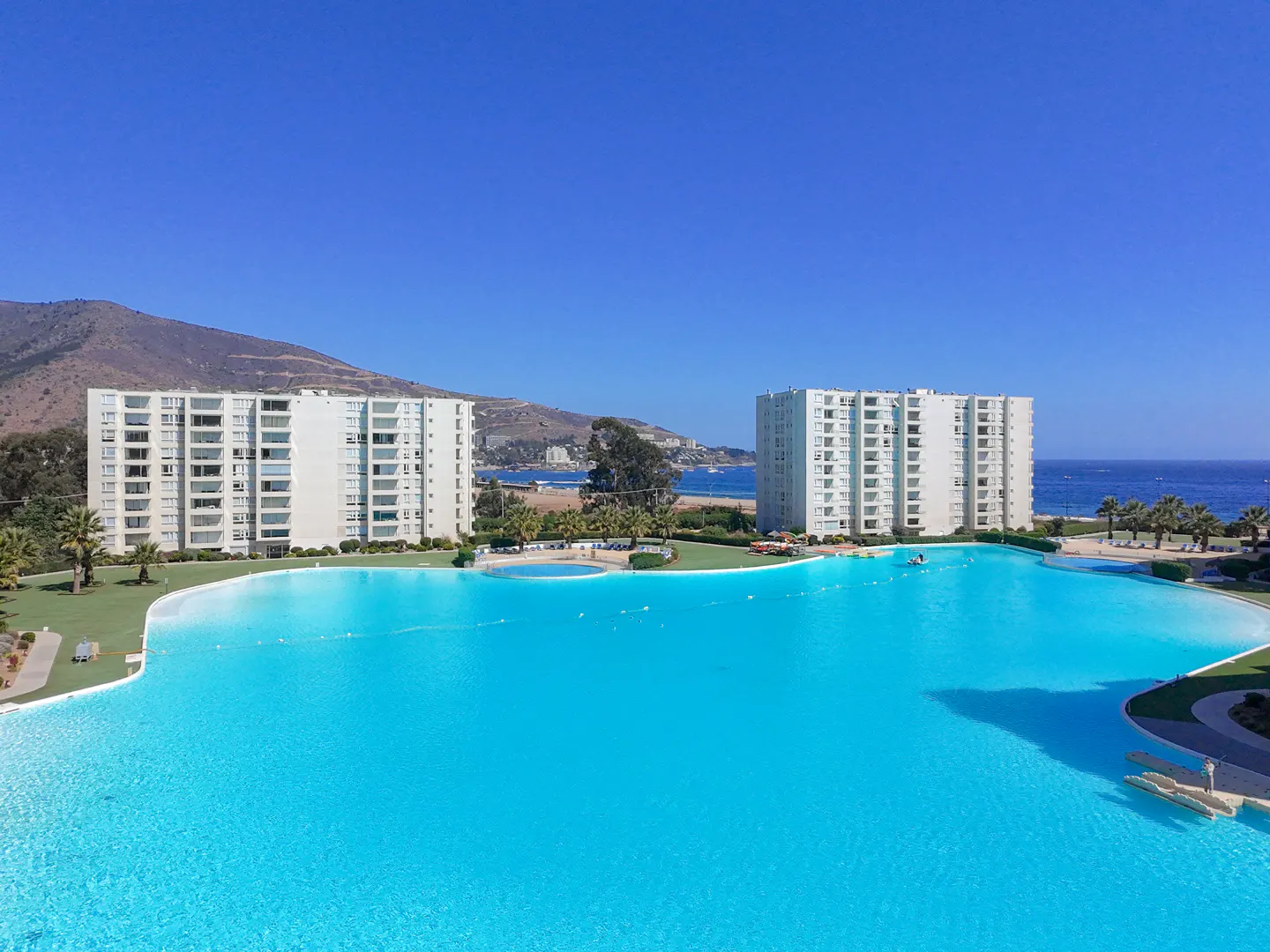 Aerial view of two white apartment buildings beside a large turquoise lagoon under a clear blue sky.