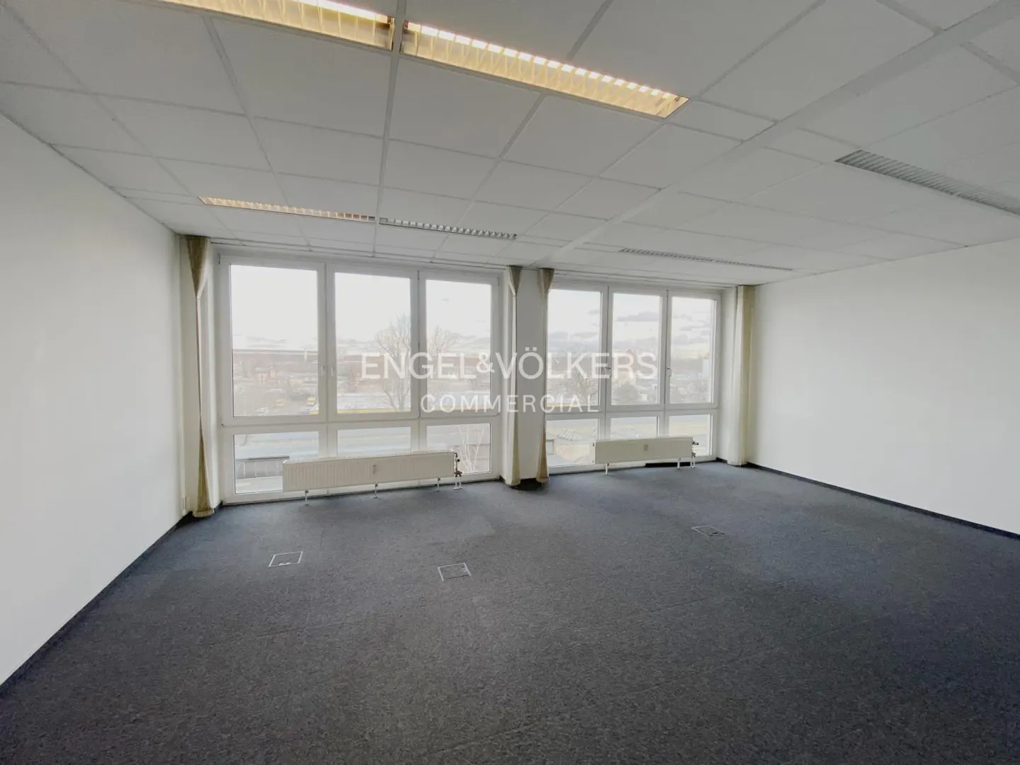 Empty office space with gray carpet, white walls, and large windows with white curtains. Engel & Völkers Commercial logo visible on the windows.