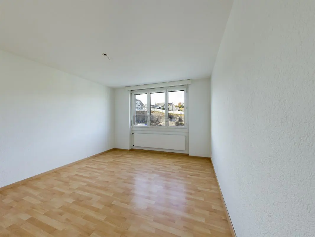 Empty room with white walls, light wood flooring, and a large window overlooking a neighborhood.