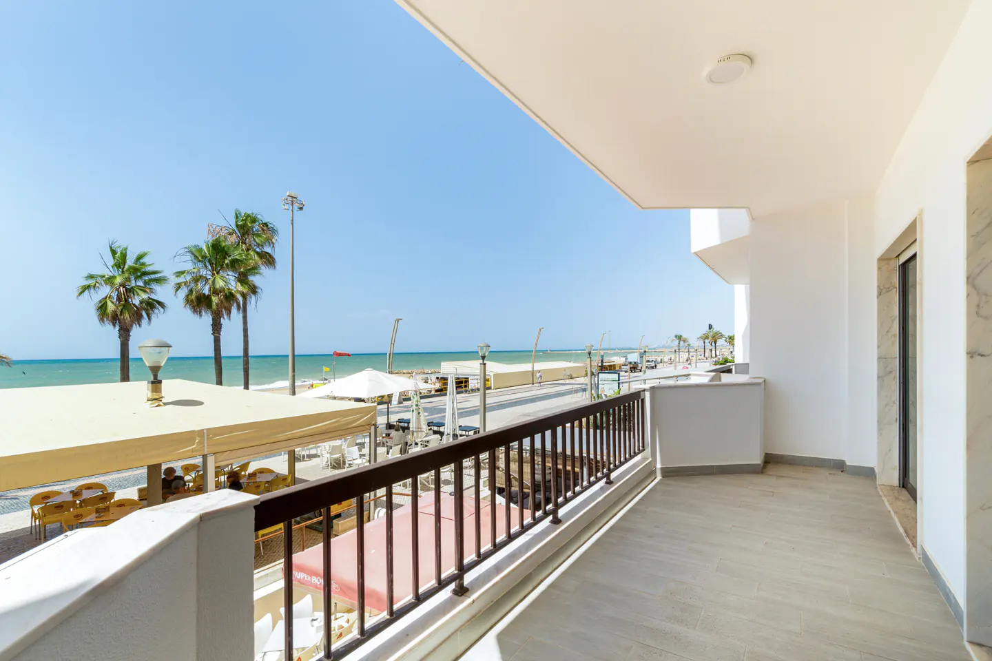Balcony view overlooking a sunny beach promenade with palm trees, outdoor dining areas, and the blue sea under a clear sky.