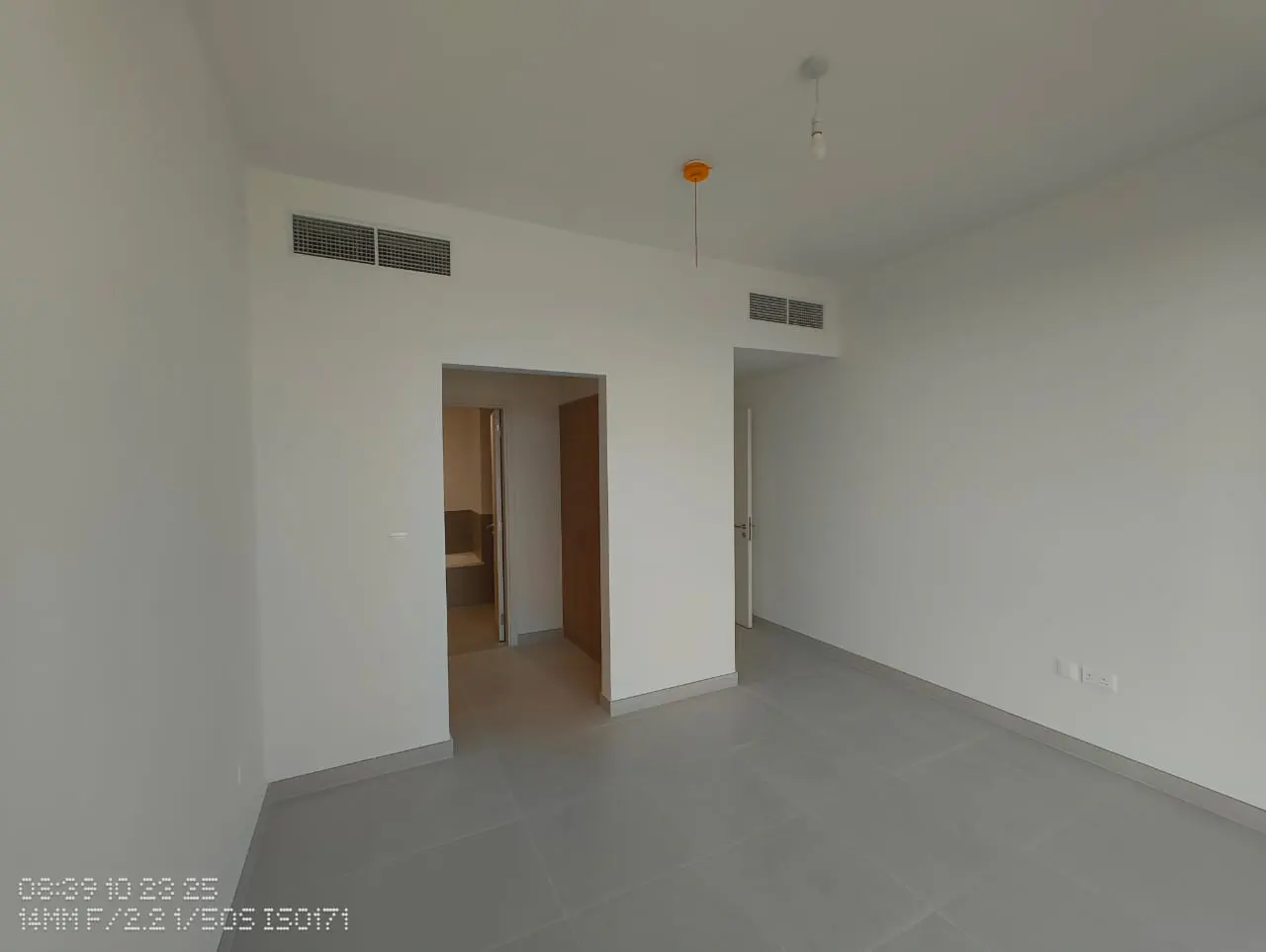 A bright, empty room with white walls, gray tile floor, and two doorways. One doorway leads to a bathroom.
