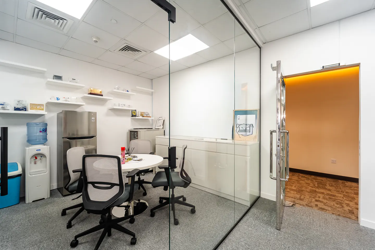 Bright office space with a glass-walled conference room, a white table, black chairs, and an open door to another room with wood floors.