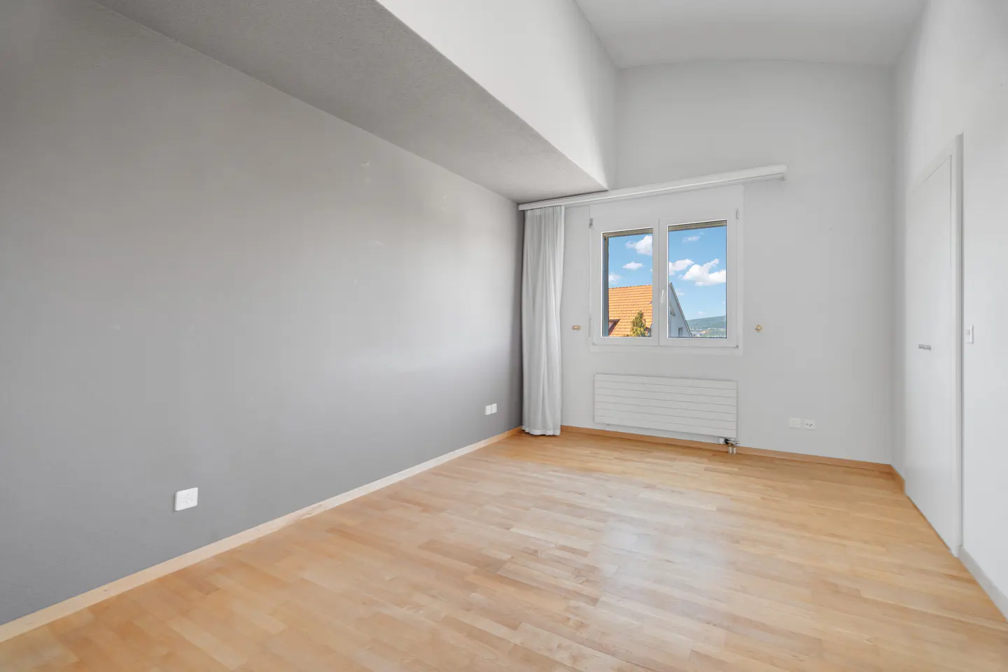 Bright, empty room with light wood floors, gray and white walls, and a window showing a blue sky and rooftops.