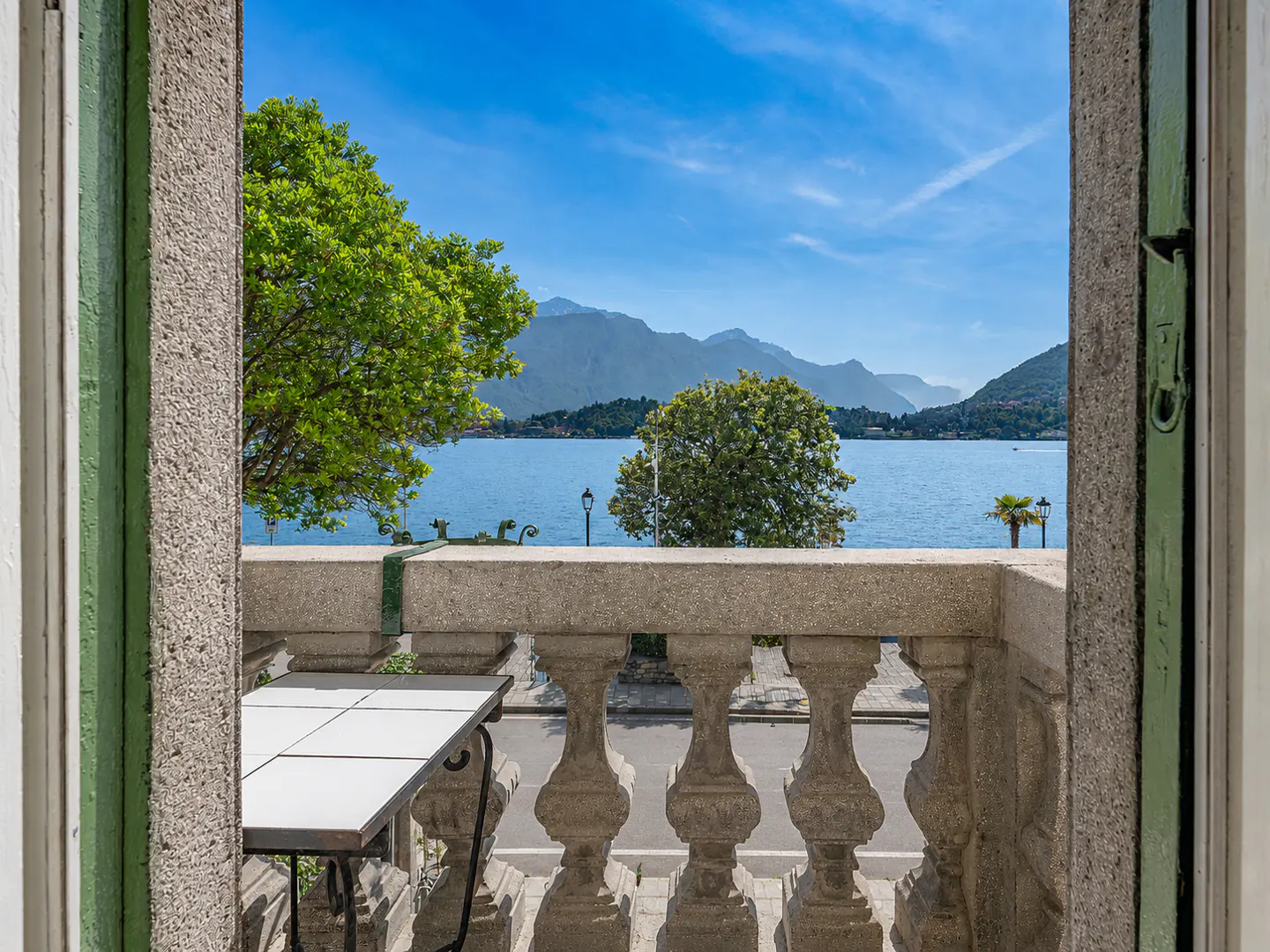 View from a balcony with a stone balustrade overlooking a blue lake and mountains under a clear sky. A table sits on the balcony.