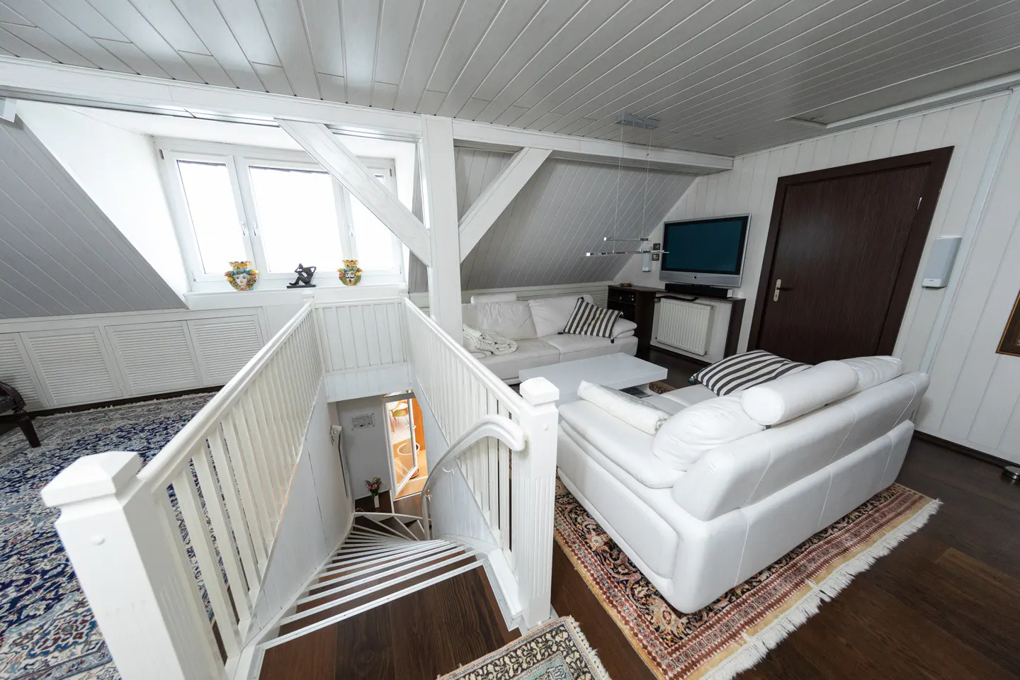 Attic living room with white walls, ceiling, and staircase. White sofas, TV, and dark wood floors. A window lets in natural light.