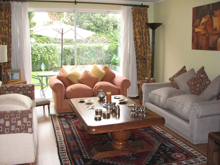 A living room with sofas, a wooden coffee table with silver objects, and a view of a garden through a sliding glass door.