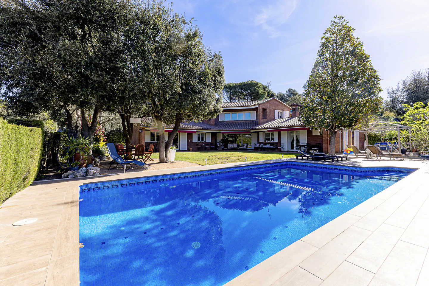A bright blue pool in front of a two-story brick house with white trim and green trees.