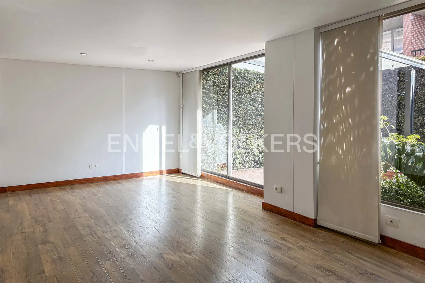 Empty room with wood floors, white walls, and large windows. Green foliage is visible outside. Natural light fills the space.