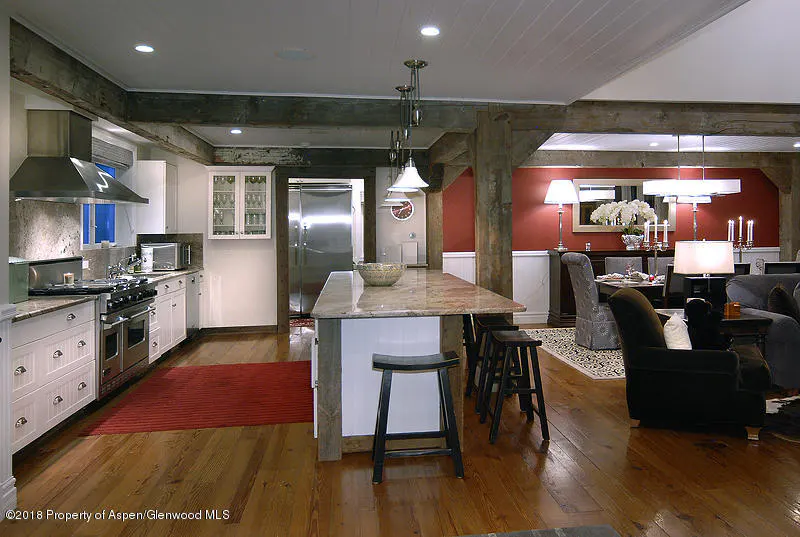 Open-concept kitchen and living area with wood floors, exposed beams, and a marble island with stools. Red accent wall in the living area.