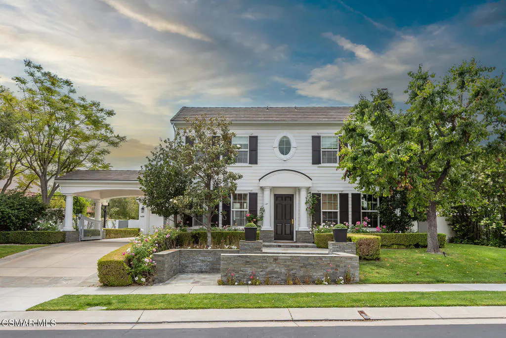 Two-story white house with black shutters, oval window, and dark door. Manicured lawn and stone retaining wall with flowers. Carport on the left.